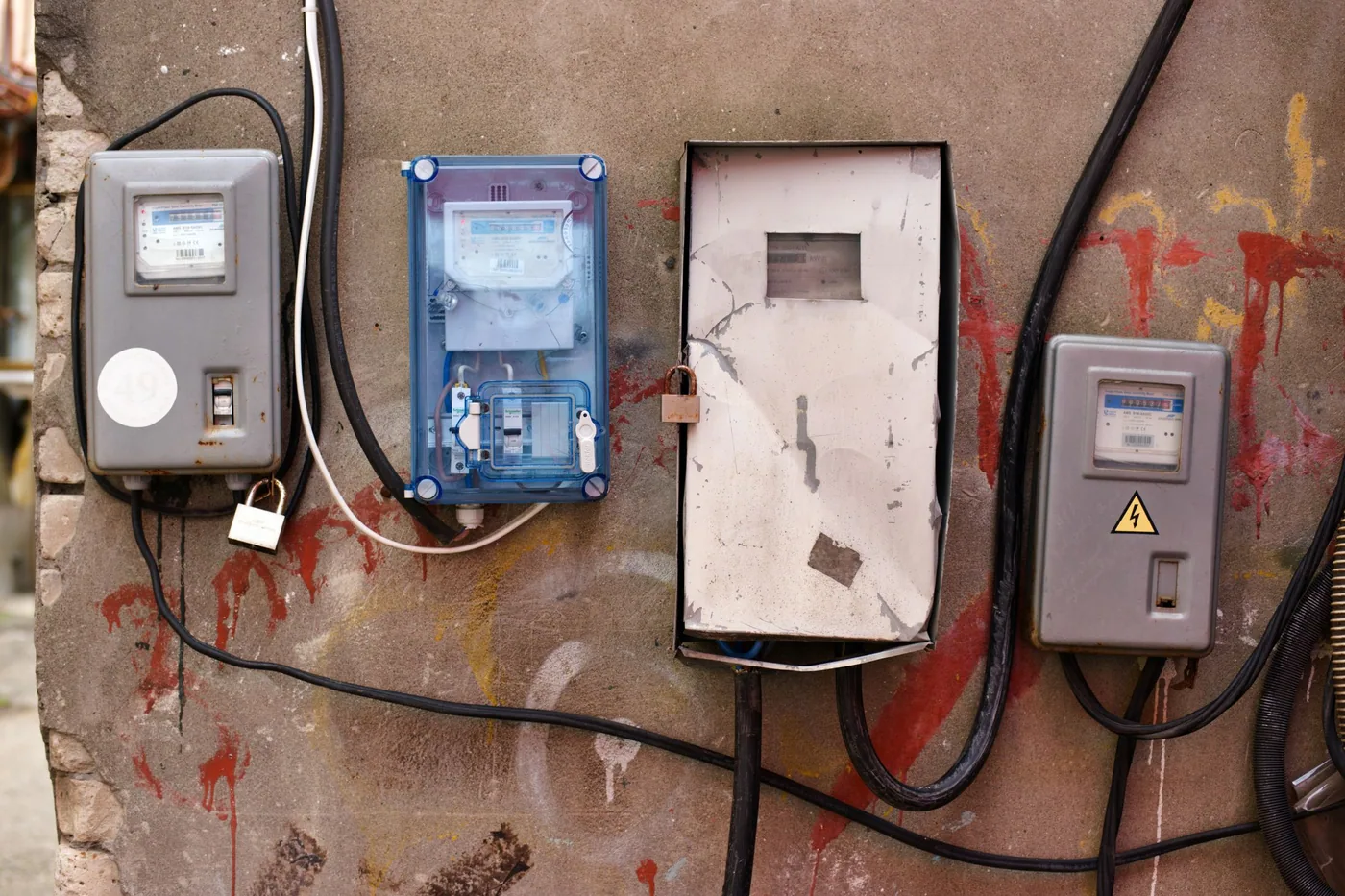 An electrician's gloved hands using a test meter inside an open distribution board, with breakers and coloured conductors visible.