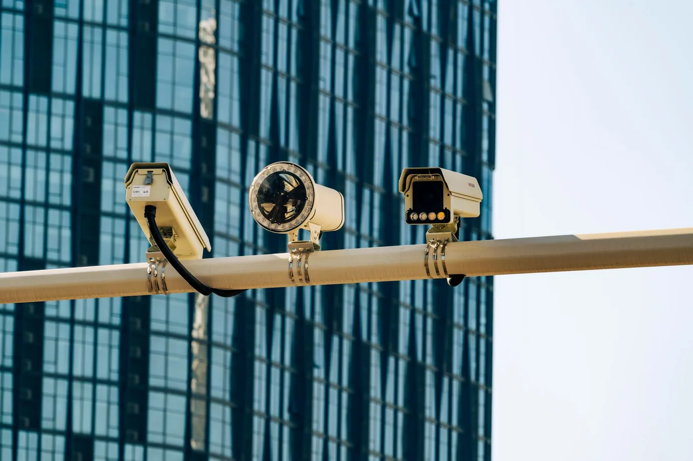 A fixed outdoor CCTV camera in a weatherproof housing mounted on a metal pole, viewed from below against a pale sky.